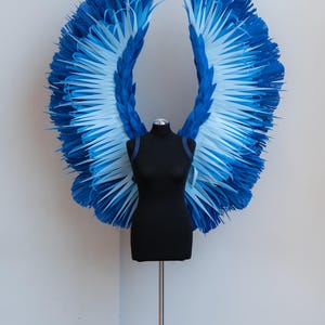 May include: A pair of large, blue and white angel wings made of paper, displayed on a black mannequin.