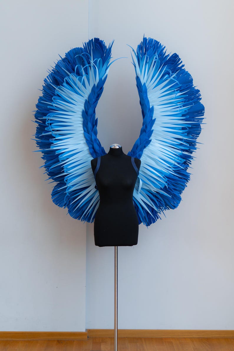 May include: A pair of large, blue and white angel wings made of paper, displayed on a black mannequin.