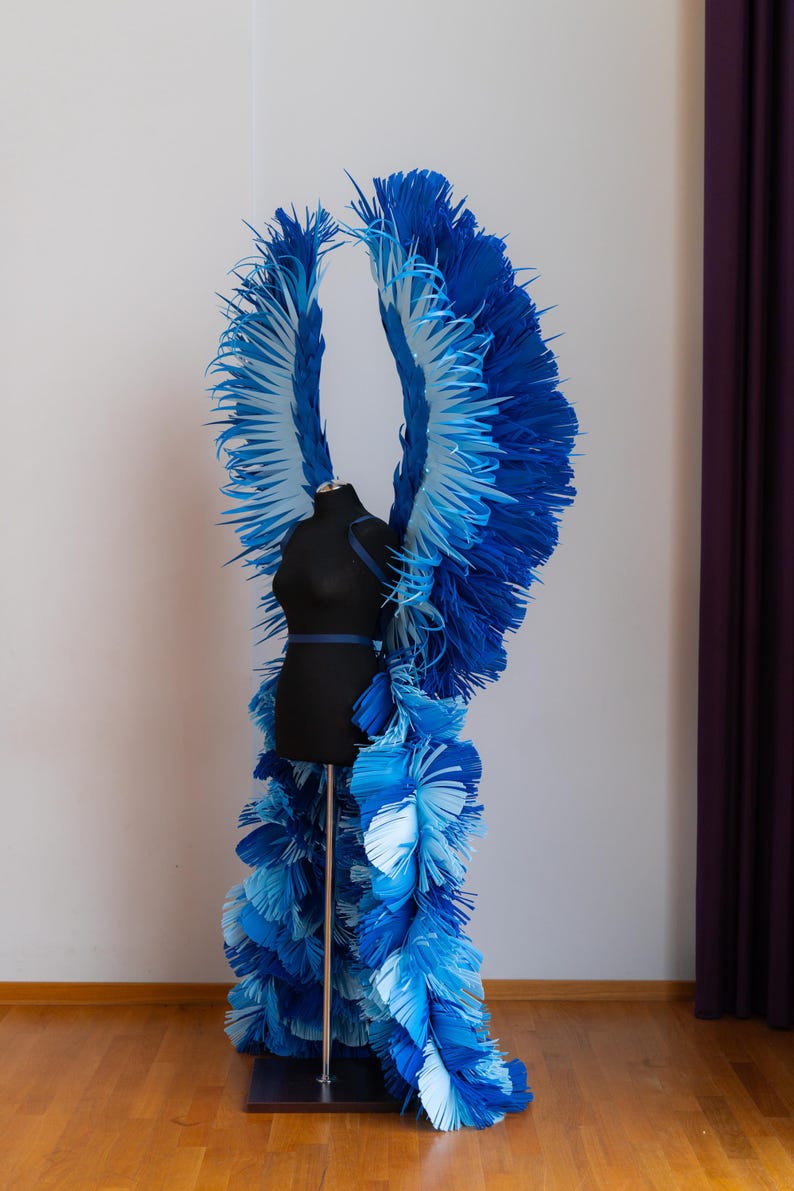 May include: A pair of large, blue and white angel wings made of paper, displayed on a black mannequin. The wings are layered and have a feathery texture.