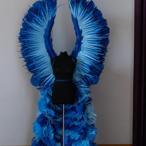 May include: A pair of large, blue angel wings with a gradient of light blue to dark blue. The wings are made of paper and are attached to a black mannequin. The wings are standing on a wooden floor.