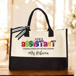 May include: A white canvas tote bag with black trim and handles. The bag has a colorful graphic that says "Admin Assistant" with flowers in the letters. The text "Professional problem solver and chaos coordinator" is below the graphic. The name "Mrs. Rebecca" is printed below the text.