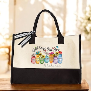 May include: A canvas tote bag with a black bottom and handles. The bag features the text "God Says You Are" and colorful illustrations of flowers in vases. The name "Victoria" is written at the bottom.