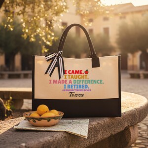 Puede incluir: Un bolso tote de lona crema con asas negras y una base negra. El bolso presenta el texto "I CAME. I TAUGHT. I MADE A DIFFERENCE. I RETIRED." con un gr&aacute;fico de manzana. El nombre "Tessa" est&aacute; impreso debajo, con un lazo a rayas blanco y negro.