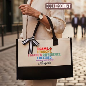 Puede incluir: Un bolso tote de lona con asas negras y un panel inferior negro. La bolsa presenta el texto "I CAME. I TAUGHT. I MADE A DIFFERENCE. I RETIRED." con un gr&aacute;fico de manzana colorida y el nombre "Angela". Un lazo a rayas blanco y negro adorna el asa.