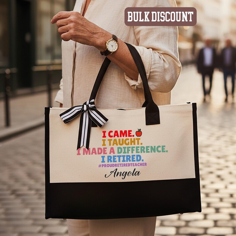 Puede incluir: Un bolso tote de lona con asas negras y un panel inferior negro. La bolsa presenta el texto "I CAME. I TAUGHT. I MADE A DIFFERENCE. I RETIRED." con un gr&aacute;fico de manzana colorida y el nombre "Angela". Un lazo a rayas blanco y negro adorna el asa.