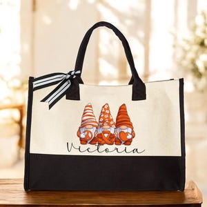 May include: A canvas tote bag with a black base and handles, featuring three gnomes with orange and white hats and holding various objects. The name "Victoria" is written in cursive below the gnomes. A black and white striped ribbon is tied to the handle.