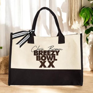 May include: A canvas tote bag with black handles and a black bottom panel. The bag features the text "Chris Brown BREEZY BOWL XX" in brown lettering. A black and white striped ribbon is tied to the side of the bag.