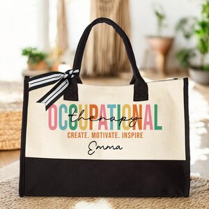 May include: A black and white canvas tote bag with a colorful "Occupational Therapy" design and the name "Emma" printed on the front. The bag has black handles and a black bottom.