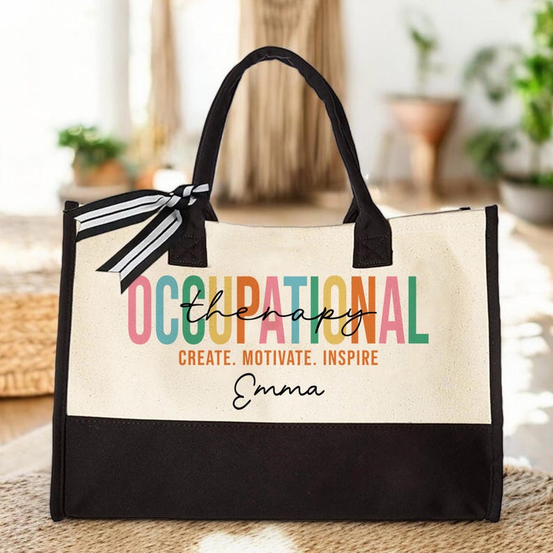 May include: A black and white canvas tote bag with a colorful "Occupational Therapy" design and the name "Emma" printed on the front. The bag has black handles and a black bottom.
