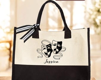 May include: A canvas tote bag with black handles and a black bottom panel. The bag features a graphic of comedy and tragedy masks with the name "Jessica" below. A black and white striped ribbon is tied to the handle.
