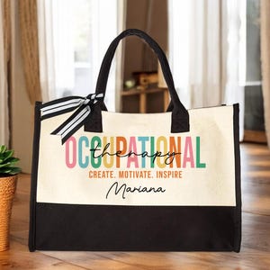 May include: A white canvas tote bag with black handles and a black bottom. The bag has a colorful design that says "Occupational Therapy" with the word "Therapy" in a smaller font. The text "Create. Motivate. Inspire" is below the design, and the name "Mariana" is written in cursive below that.