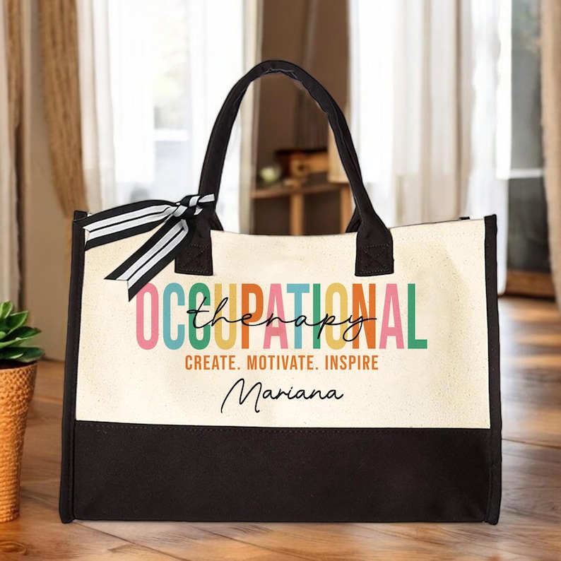 May include: A white canvas tote bag with black handles and a black bottom. The bag has a colorful design that says "Occupational Therapy" with the word "Therapy" in a smaller font. The text "Create. Motivate. Inspire" is below the design, and the name "Mariana" is written in cursive below that.