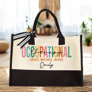 May include: A beige canvas tote bag with black handles and a black bottom. The bag has a colorful design that says "Occupational Therapy" with the word "Therapy" in a smaller font. The words "Create. Motivate. Inspire" are printed below the design. The name "Emily" is written in cursive below the words.