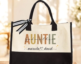 May include: A beige and black tote bag with the text "AUNTIE Godmother marcela david" in colorful letters. The bag has black handles and a black and white striped ribbon.