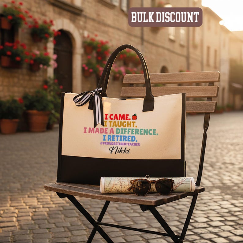 Puede incluir: Bolso tote color crema con asas y base negras. El bolso muestra el texto "I CAME. I TAUGHT. I MADE A DIFFERENCE. I RETIRED." con un gr&aacute;fico de manzana. Un lazo a rayas blanco y negro est&aacute; atado al asa.
