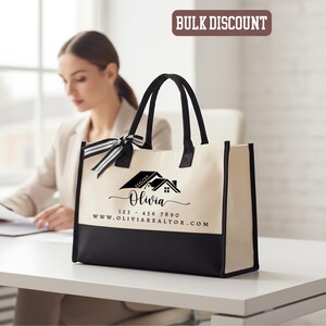 May include: Cream-colored tote bag with black accents and handles. The bag features a black bottom panel and a black and white striped bow. The text "Olivia" and a house logo are printed on the front, along with a phone number and website.
