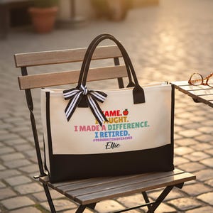 Puede incluir: Bolso tote color crema con asas negras y un panel inferior negro. El bolso presenta un lazo a rayas blanco y negro y el texto: "Vine, ense&ntilde;&eacute;, marqu&eacute; la diferencia, me jubil&eacute;". El nombre "Ellie" est&aacute; impreso debajo.