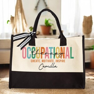 May include: A white canvas tote bag with black handles and a black bottom. The bag has a colorful design that says "Occupational Therapy" with the word "Therapy" written in a smaller font. The bag also has the text "Create. Motivate. Inspire" and the name "Camilla" written in black.