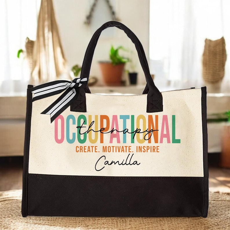 May include: A white canvas tote bag with black handles and a black bottom. The bag has a colorful design that says "Occupational Therapy" with the word "Therapy" written in a smaller font. The bag also has the text "Create. Motivate. Inspire" and the name "Camilla" written in black.