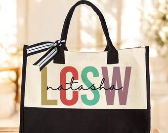 May include: A white canvas tote bag with black handles and a black bottom. The bag has the letters "LCSW" printed in colorful block letters with the name "natasha" written in black script underneath. The bag has a black and white striped ribbon tied to the handles.