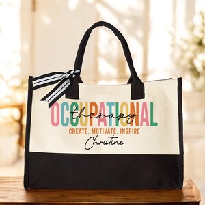 May include: A white canvas tote bag with black handles and a black bottom. The bag has a colorful graphic that says "Occupational Therapy" with the word "therapy" in a smaller font. The text "Create. Motivate. Inspire" is below the graphic. The name "Christine" is written in cursive below the text.