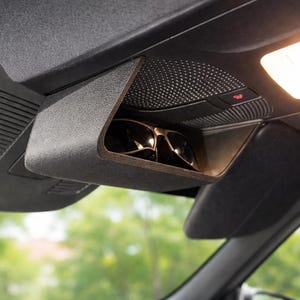 Op de afbeelding: Een close-up van het interieur van een auto, met een ingebouwde zonnebrilhouder. De houder is donkergrijs en bevat een zonnebril. Bovenin schijnt een lamp. De foto is genomen vanuit een lage hoek.