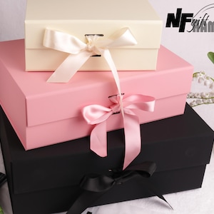 Op de afbeelding: Drie gestapelde geschenkdozen in crème, roze en zwart, elk vastgebonden met een satijnen strik. De bovenste doos is crème, de middelste doos is roze en de onderste doos is zwart. De dozen zijn in een piramidevorm gerangschikt.