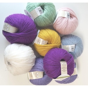 May include: A collection of eight yarn balls in various colors including purple, white, light green, pale pink, mustard yellow, and light blue. Each ball features a white paper label with printed text and a barcode wrapped around the center.