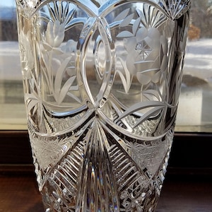 May include: A clear, cut-glass vase with a wide, scalloped rim. The vase features etched floral and geometric patterns, with a flared base and a tall, cylindrical body. The intricate designs catch the light, creating a sparkling effect. The vase is likely made of crystal.