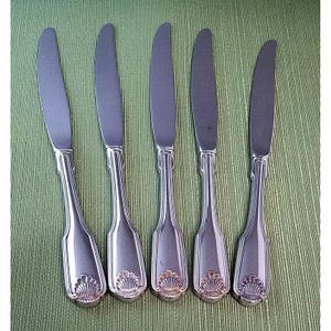 May include: A set of five silver-toned metal dinner knives arranged side by side. Each knife features a smooth, tapered blade and a handle decorated with a raised, fan-shaped shell motif at the base, complemented by vertical grooved detailing.