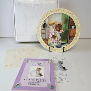 May include: A decorative plate featuring a scene from "The Tale of Two Bad Mice" by Beatrix Potter. The plate shows a mouse holding a broom. The image includes a book, a box, and other related documents. The plate is displayed on a black stand.