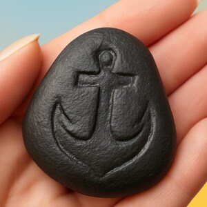Puede incluir: Una piedra negra con un ancla tallada en ella. La piedra está siendo sostenida en una mano.