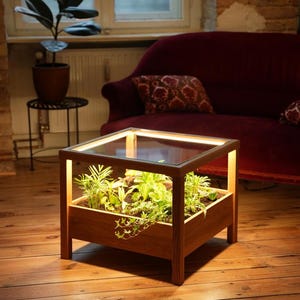 Puede incluir: Una mesa de madera con una tapa de vidrio y una maceta integrada. La maceta está llena de plantas verdes y tiene una luz integrada. La mesa está iluminada desde adentro, proyectando un brillo cálido.
