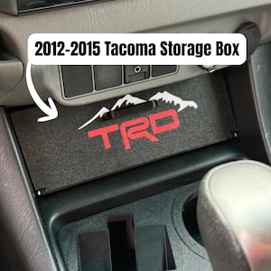 May include: Black storage box for a 2012-2015 Tacoma. The box features a red "TRD" logo and a white mountain range design. The box is designed to fit in the centre console of the vehicle.