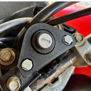 May include: Close-up of a black motorcycle ignition switch with a silver keyhole and the word "IGNITION" printed on the switch. The switch is mounted on a black metal frame with two silver screws.