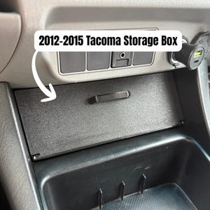 May include: A black storage box for a 2012-2015 Tacoma. The rectangular box has a small handle and fits into the vehicle's dashboard. The text "2012-2015 Tacoma Storage Box" is in a white box above the storage compartment.