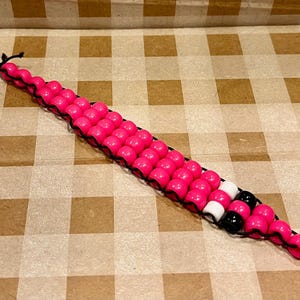 May include: A pink and black beaded keychain with a black string. The keychain is made of small, round beads and has a black string attached to it.
