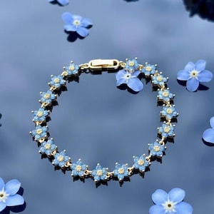 May include: A gold-toned bracelet featuring blue flower-shaped charms. The bracelet is adorned with small, light blue flowers with yellow centers. Scattered around the bracelet are several blue forget-me-not flowers, creating a delicate and floral aesthetic.