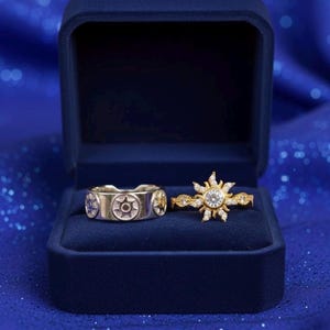 May include: Two rings displayed in a blue velvet box. One ring is silver with a repeating sunburst design. The other is gold with a central clear stone and sunburst design. The box is open, revealing the rings against a blue background.