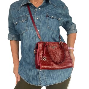 May include: A red leather handbag with a crocodile embossed pattern and a gold heart charm. The bag has a top handle and a long shoulder strap.
