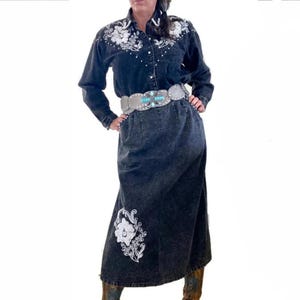 May include: A black denim skirt with white embroidered floral designs. The skirt has a high waist and a long, flowing silhouette. A silver belt with turquoise stones is worn around the waist.