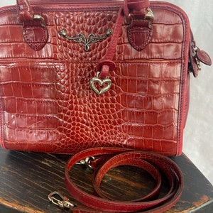 May include: A red leather handbag with a crocodile embossed pattern. The bag has a top handle and a detachable shoulder strap. The bag has a heart-shaped charm attached to it.