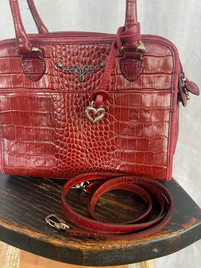 May include: A red leather handbag with a crocodile embossed pattern. The bag has a top handle and a detachable shoulder strap. The bag has a heart-shaped charm attached to it.