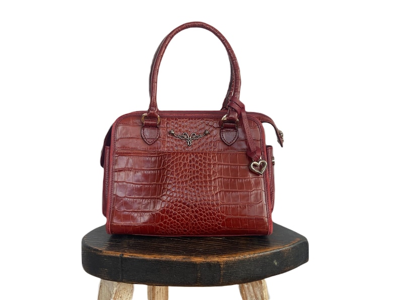 May include: A red leather handbag with a crocodile pattern and a heart-shaped charm. The bag has a top handle and a zipper closure.