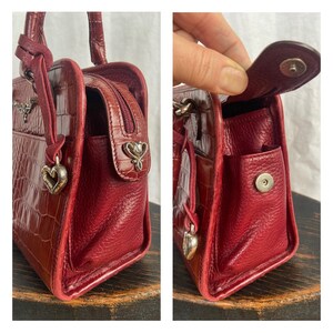 May include: A red leather handbag with a crocodile pattern. The bag has a top handle, a zipper closure, and a small silver charm hanging from the handle. The bag is sitting on a wooden surface.