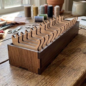 Op de afbeelding: Een donkerhouten doos met meerdere lichtbruine katvormige draad houders. De doos staat op een houten tafel, met naai benodigdheden en een raam op de achtergrond. De katvormige houders zijn voor het naaien.