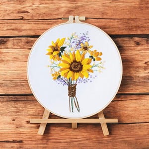 May include: A white embroidery hoop with a floral design of sunflowers and purple and white flowers. The hoop is on a wooden easel.