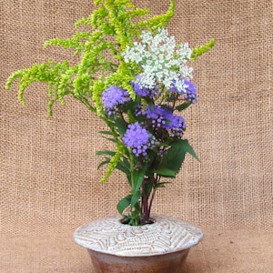 May include: A ceramic vase with a wide, patterned rim holds a vibrant floral arrangement. The arrangement features green, white, and purple flowers. The vase has a brown base and a cream-colored top. The background is a textured, tan fabric.