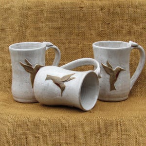 May include: Three off-white ceramic mugs with a speckled finish, each featuring a raised, textured relief of a hummingbird in flight. The mugs have a cylindrical body, a curved handle, and a small bird-shaped accent on the top of the handle.