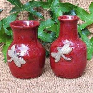 May include: Two red ceramic vases with a dragonfly design. The vases have a rounded shape and a textured surface. The dragonfly is a light beige color and is attached to the front of each vase. The vases are displayed on a burlap surface.